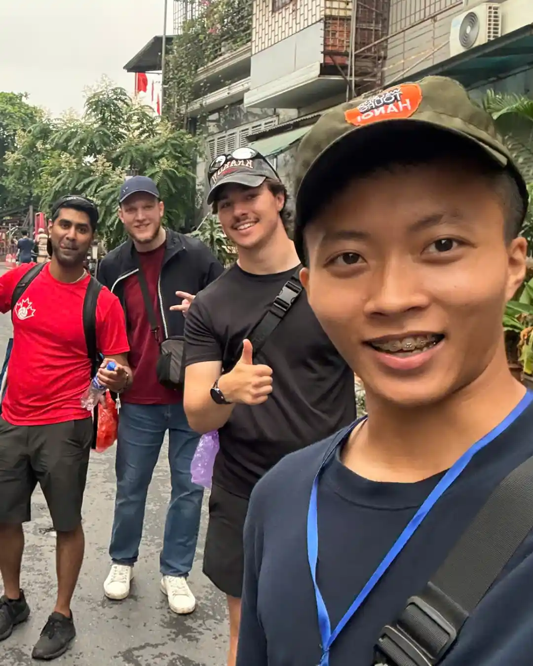 Local Hanoi guide in a green Free Walking Tours Hanoi cap leading three male travellers down a Hanoi residential street, one giving a thumbs up to the camera.