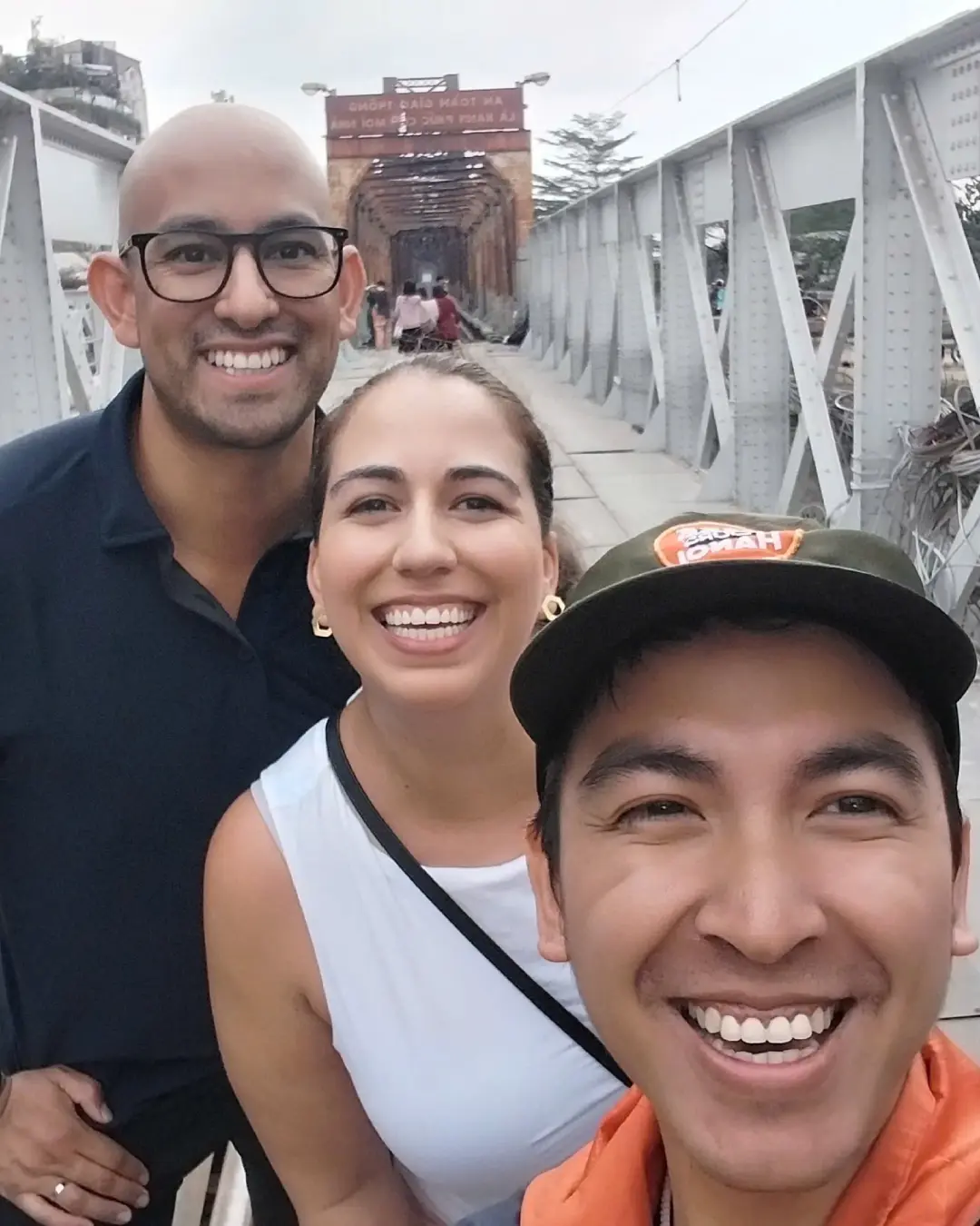 Local Hanoi guide in a Free Walking Tours Hanoi cap taking a selfie with two travellers at the entrance of Long Bien Bridge, looking down the long iron-truss pedestrian walkway over the Red River.