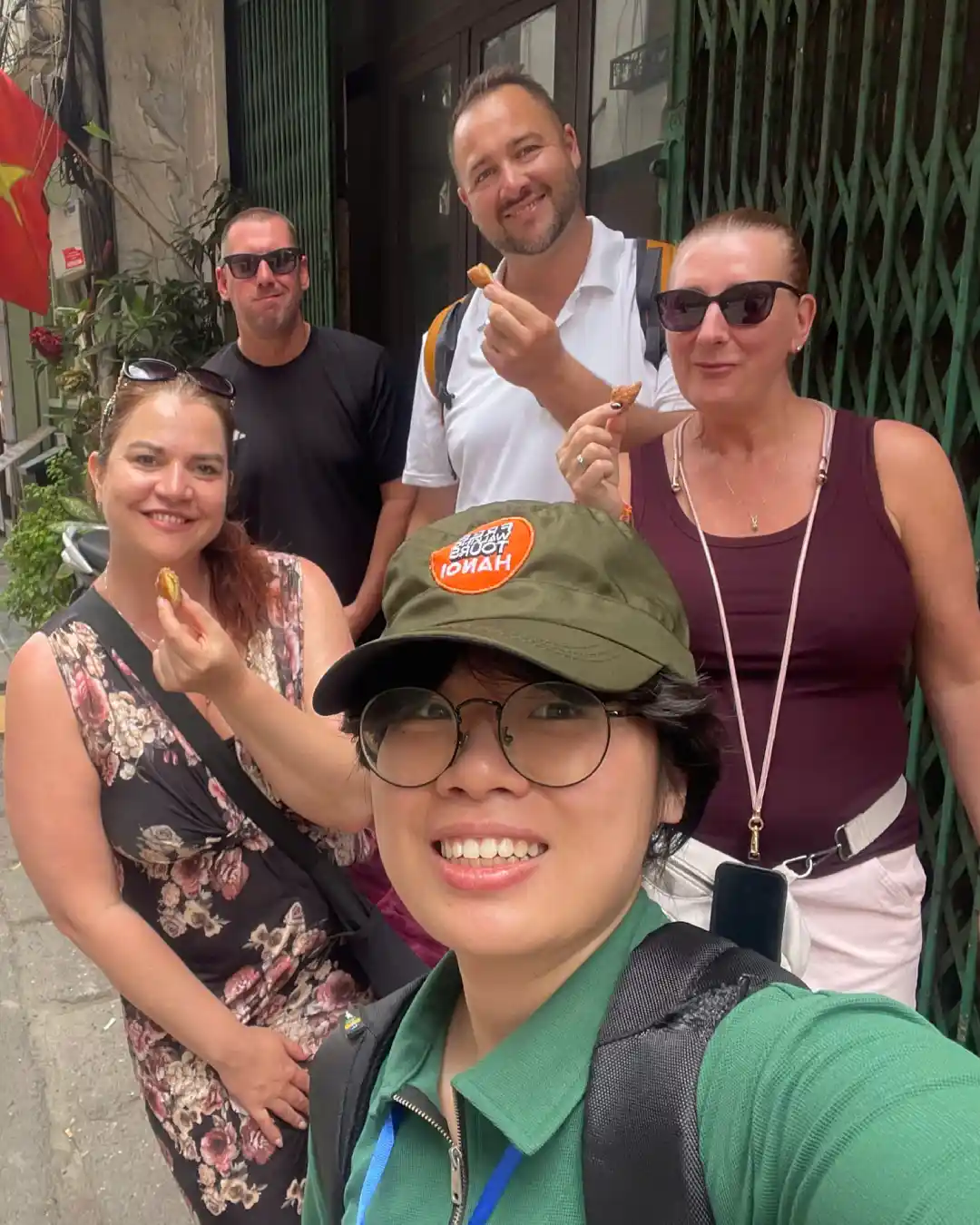 Local Hanoi guide in a Free Walking Tours Hanoi cap taking a selfie with four travellers eating fried street snacks in a narrow Old Quarter alley with a Vietnamese flag overhead.