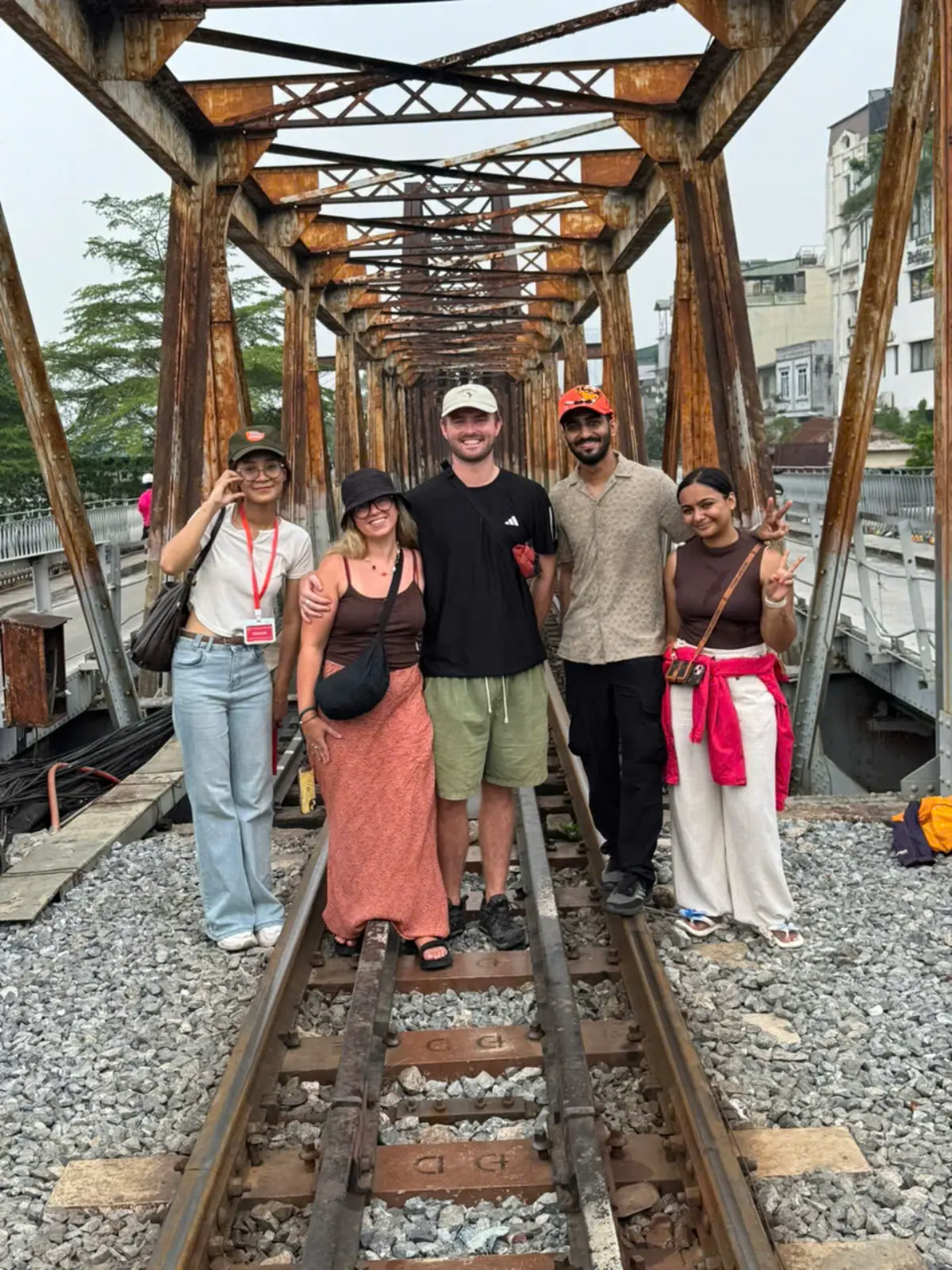 Five travellers and guide Hannah posing together on the rusted railway tracks of Long Bien Bridge in late afternoon light, framed by the bridge's century-old iron trusses.